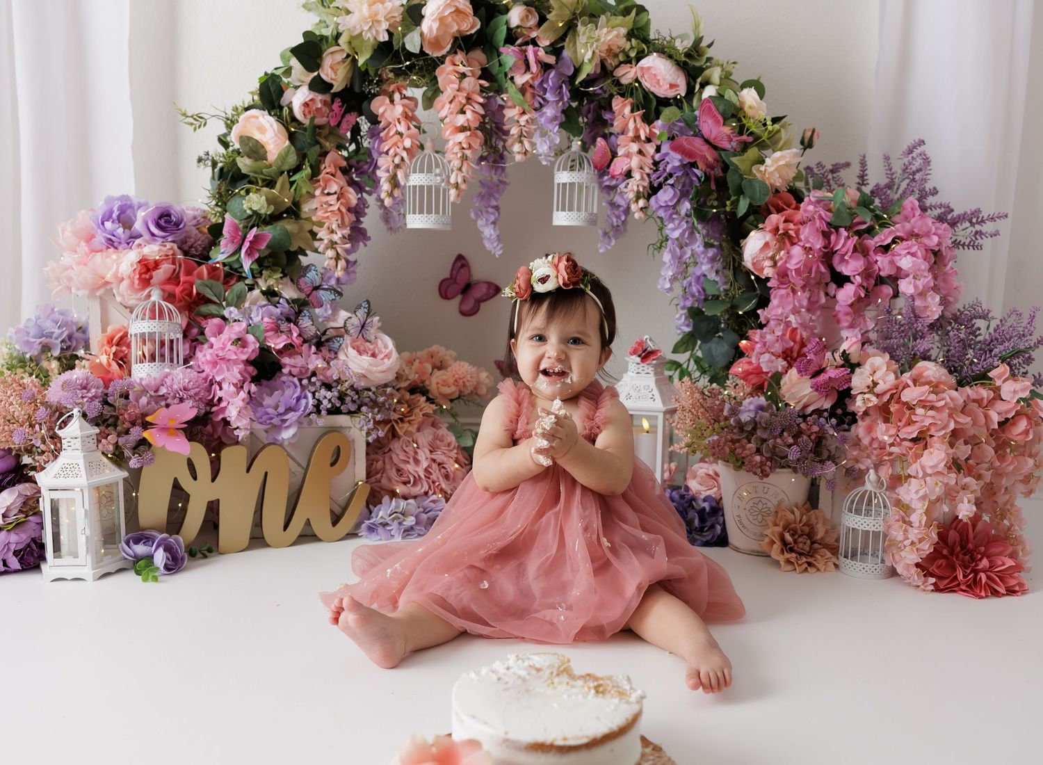 Baby girl in pink dress sits before floral arch with birdcages celebrating her first birthday cake smash.