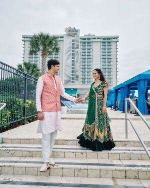 Indian couple in traditional attire holding hands on steps with a beachfront hotel in the background.