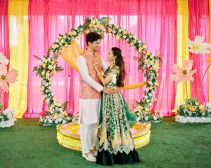 Couple in traditional Indian attire poses under floral arch with pink and yellow backdrop at wedding ceremony.