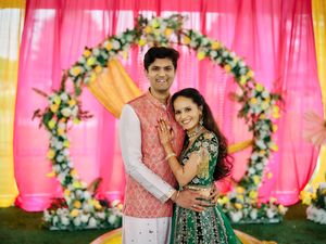 Couple posing in traditional Indian wedding attire before a floral arch with pink and yellow backdrop.