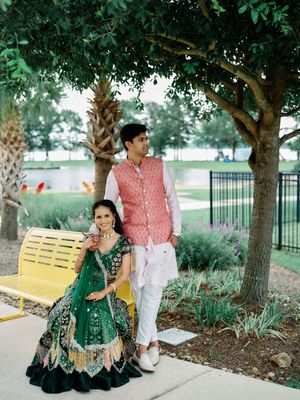 Couple in traditional Indian attire poses outdoors, woman in green lehenga seated on yellow bench, man in pink jacket standing.
