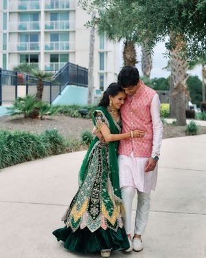 Couple in traditional Indian wedding attire, woman in green lehenga, man in pink sherwani, sharing a romantic moment outdoors.