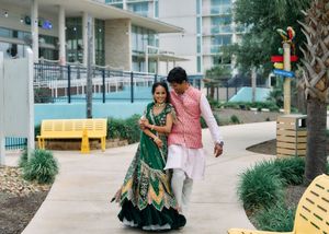 Indian couple in traditional attire walking together outdoors, bride in green lehenga, groom in pink sherwani.