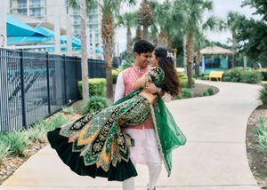 Groom lifting bride in green embroidered lehenga on a scenic outdoor pathway with palm trees.