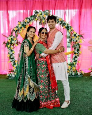 Indian wedding trio posing under a floral arch with vibrant pink backdrop during a festive celebration.