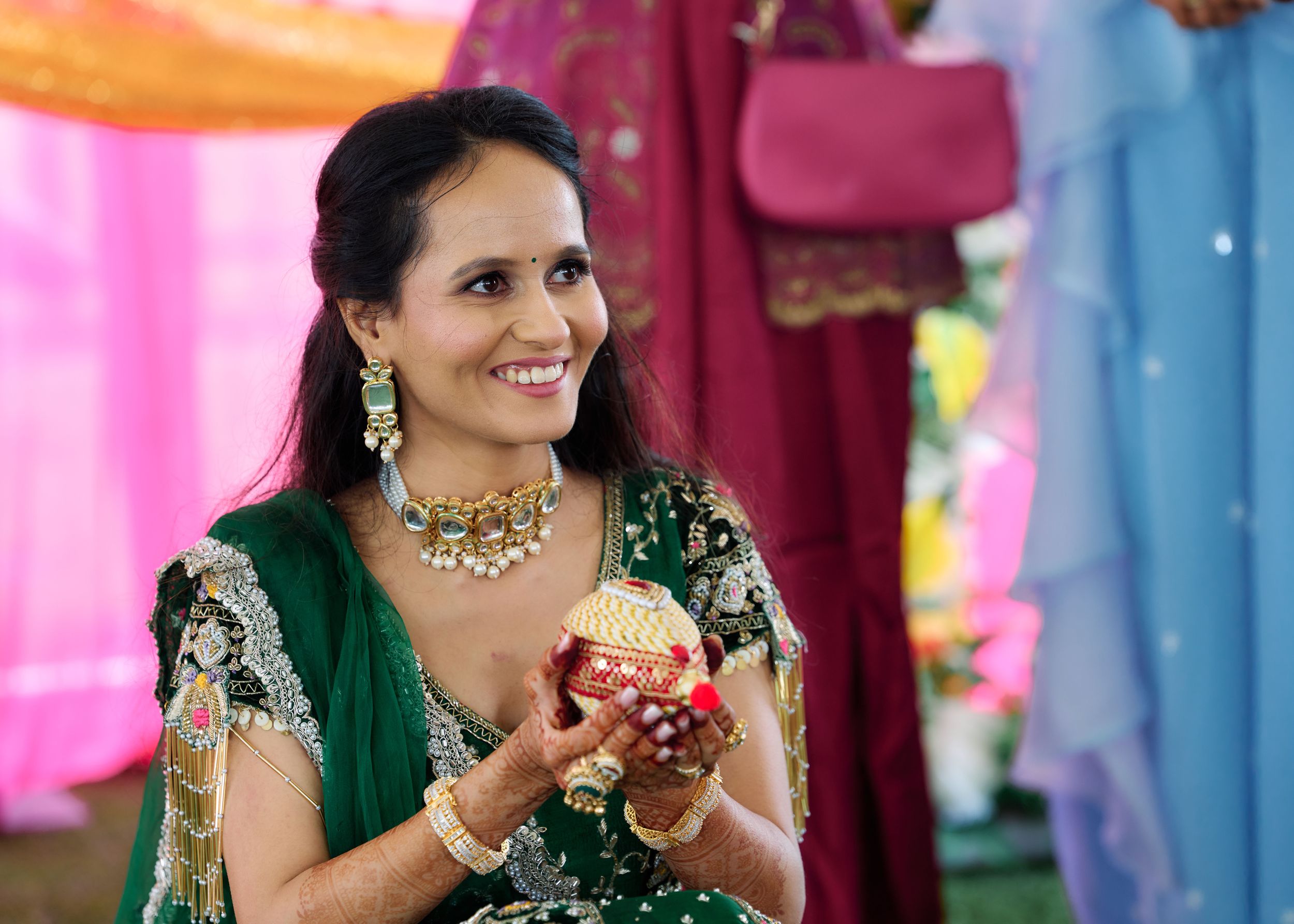 Smiling Indian woman in green embroidered lehenga holding decorative pot at traditional ceremony.