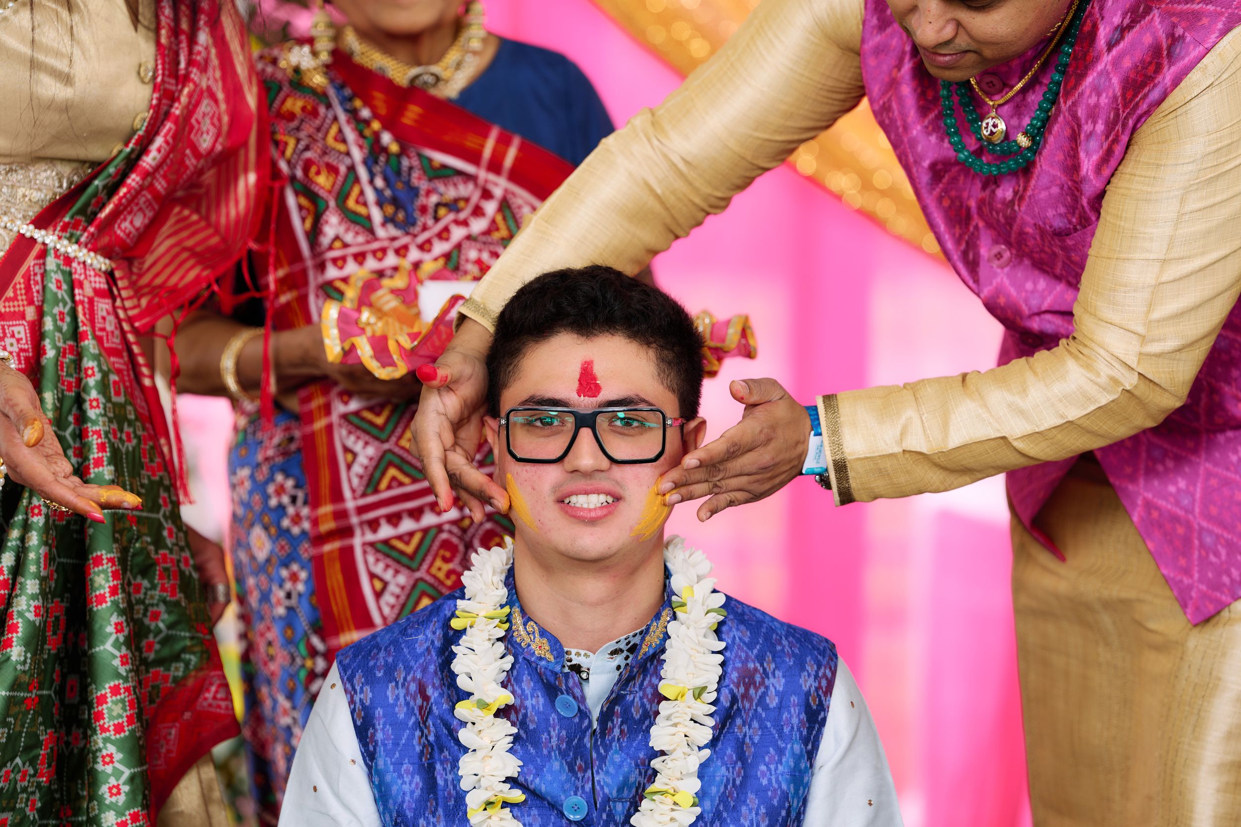 Young man in blue vest and glasses receives tilak blessing during Indian wedding ceremony with flower garland.
