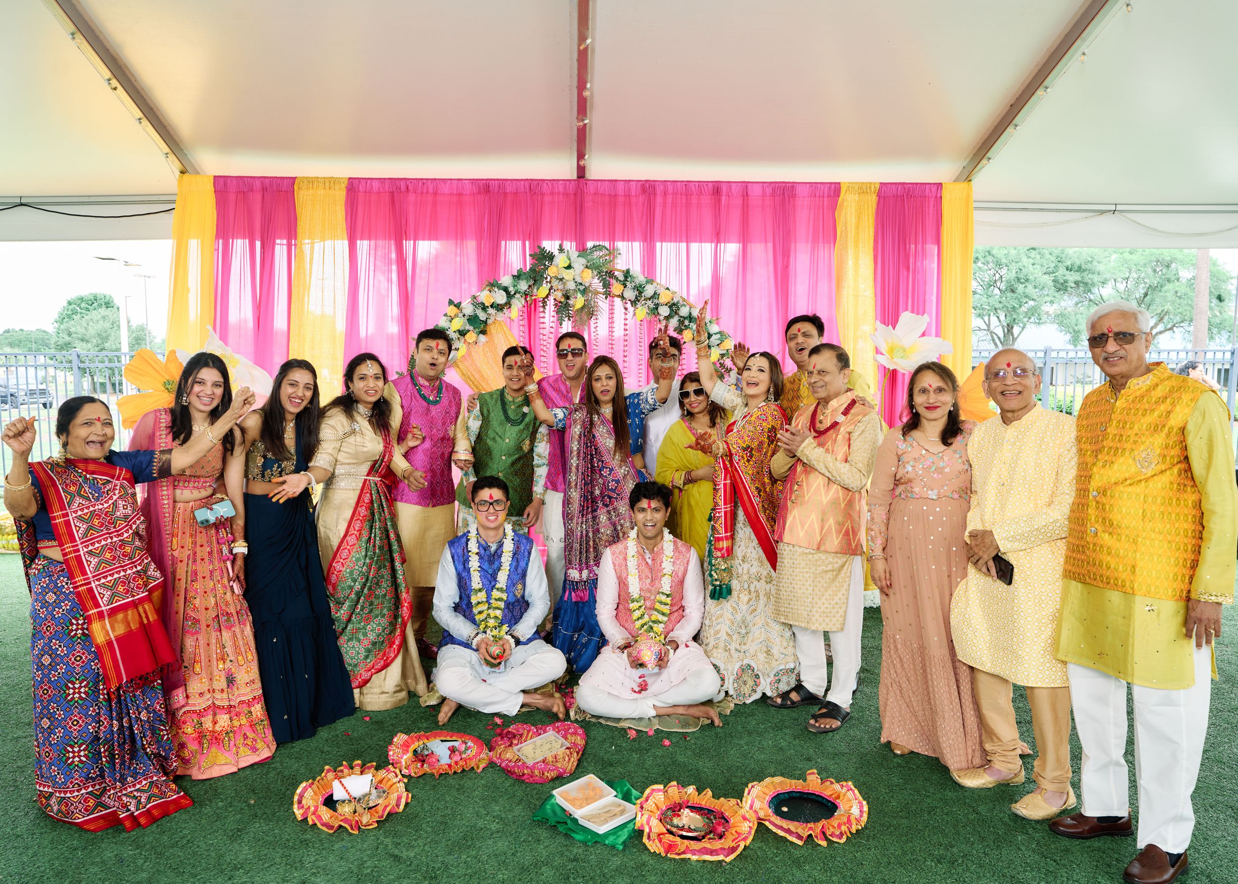 Vibrant Indian wedding celebration with family and friends posing under a colorful floral mandap with pink and yellow decor.