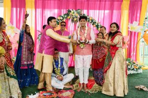 Colorful Indian wedding ceremony with groom in white, guests in vibrant traditional attire under a floral mandap.