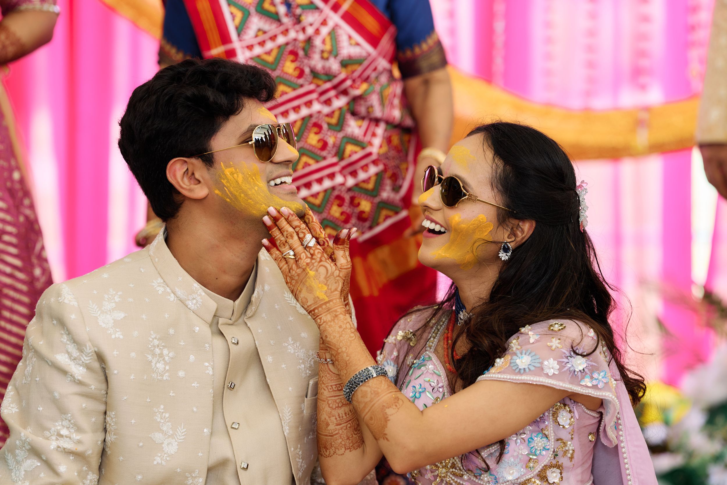 Couple laughing during Indian Haldi ceremony, wearing sunglasses with turmeric on faces, colorful backdrop.