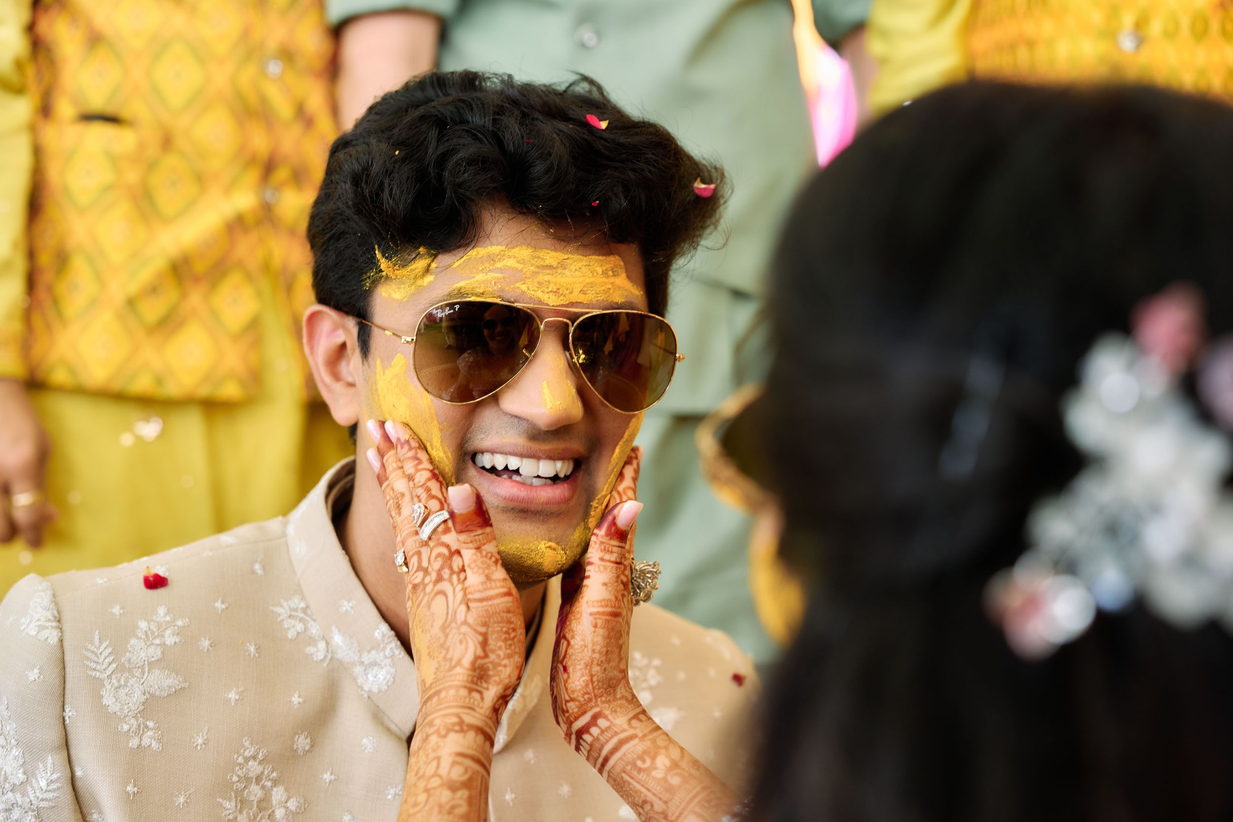 Groom smiling with turmeric haldi on face during Indian wedding ceremony, wearing sunglasses and beige sherwani.