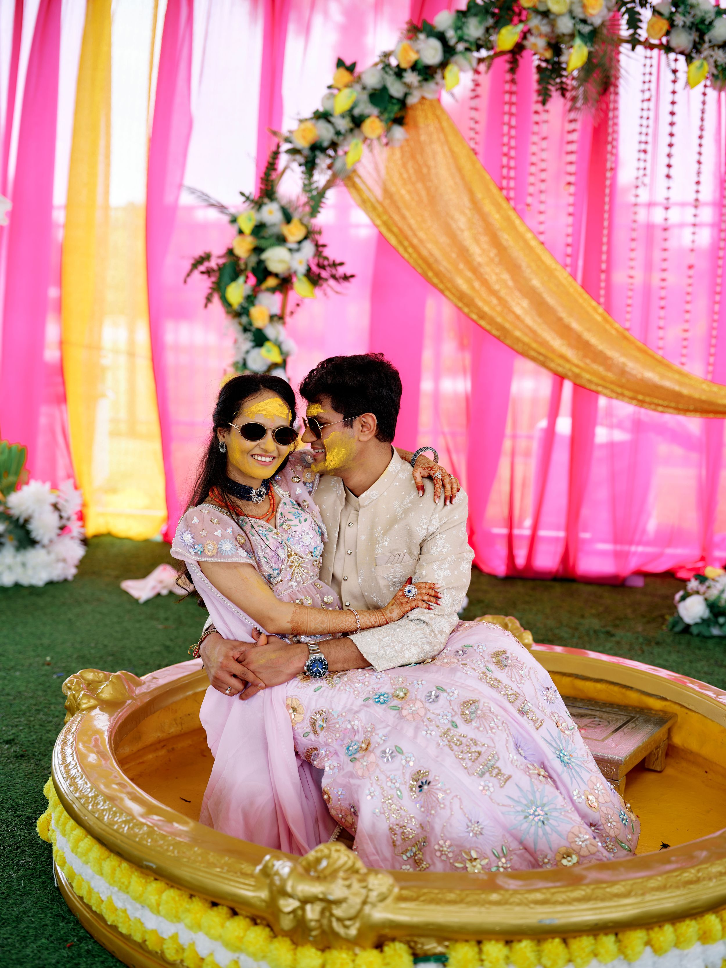 Indian couple sitting together in a decorative golden seat during a colorful Haldi wedding ceremony.