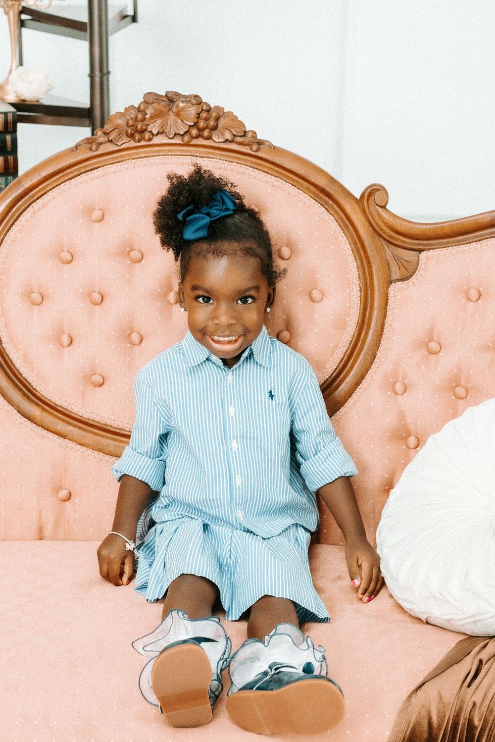 Young girl in Ralph Lauren striped dress and silver shoes smiles on pink tufted vintage chair.