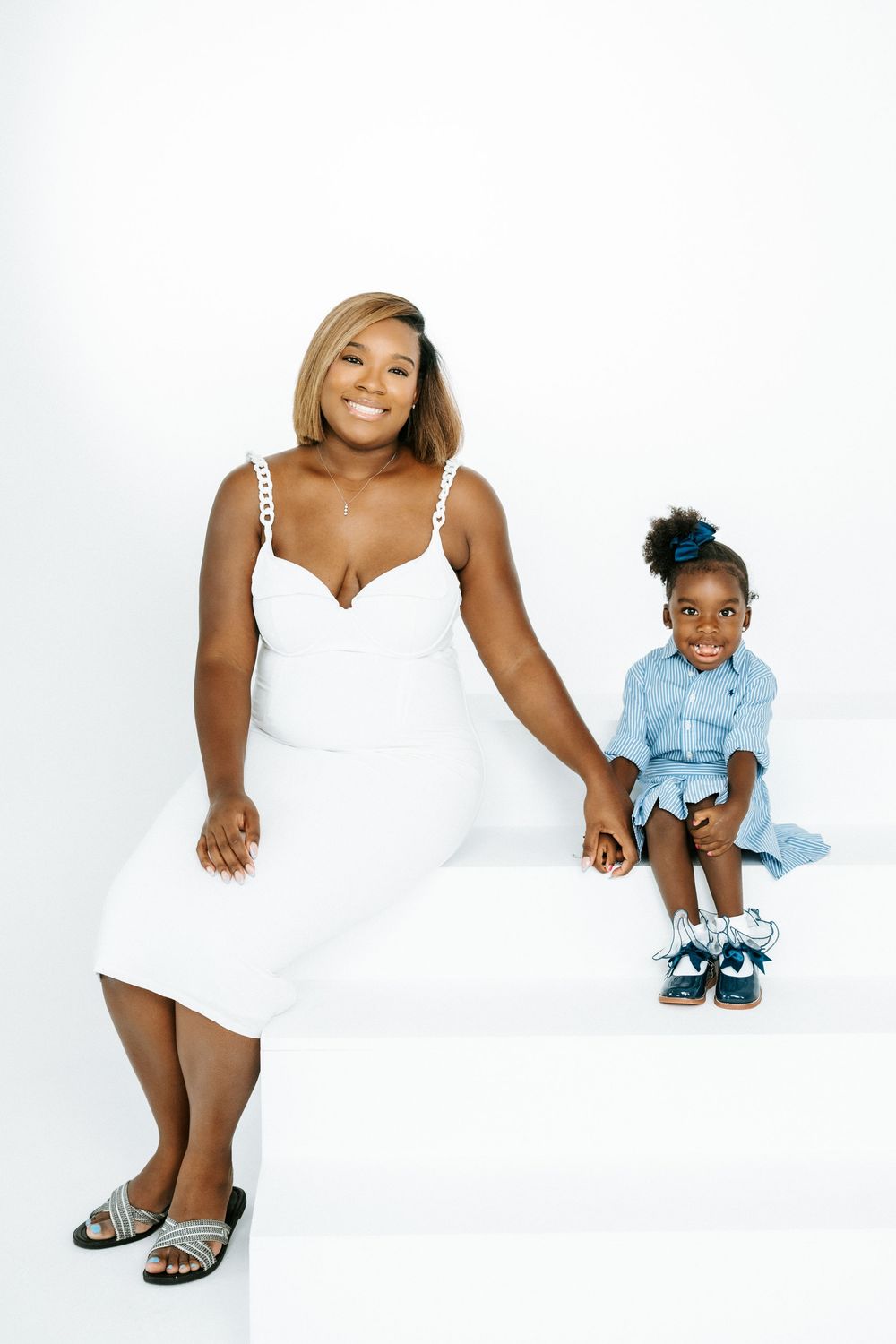 A woman in a white dress sits with a young girl in a blue outfit on a white backdrop.
