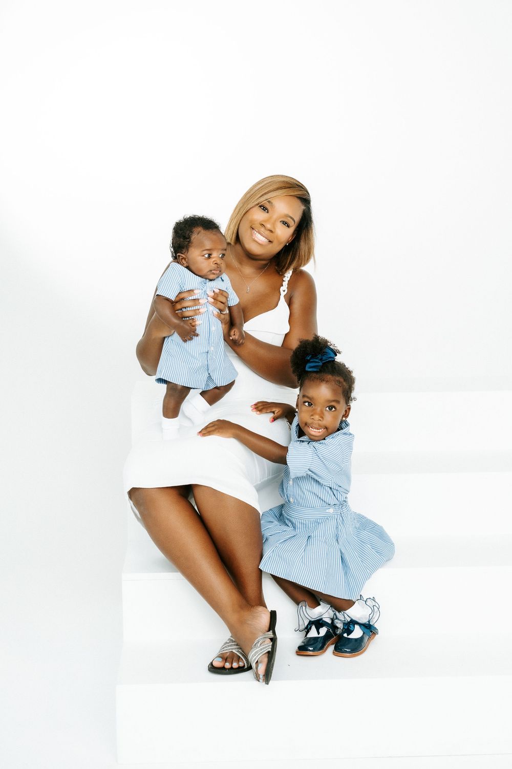A smiling mother in a white dress sits on white steps holding her baby while her toddler daughter stands beside her.