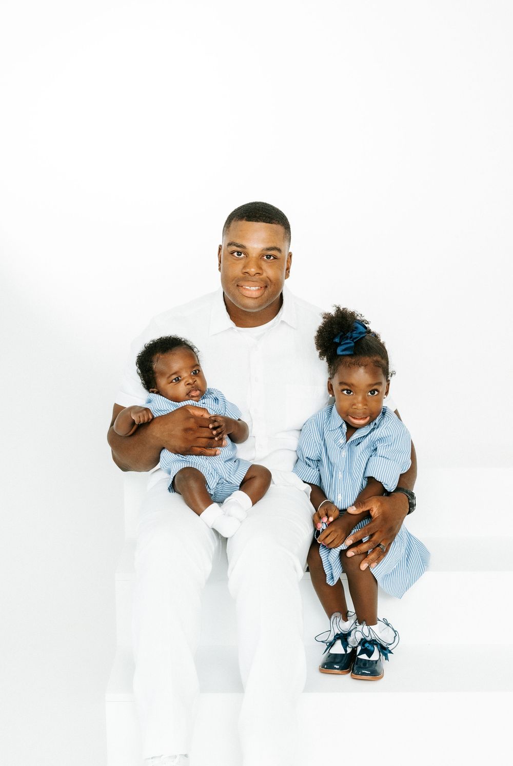 A father in white holds his baby and toddler daughter, both in blue outfits, against a white background.