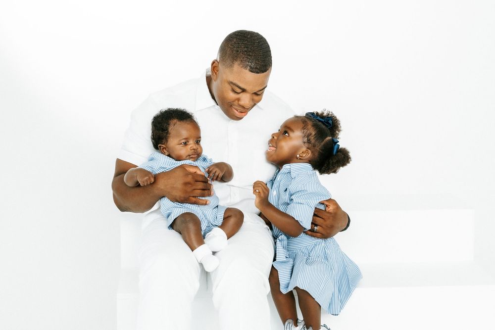 A father in white holds his baby and toddler daughter in blue outfits against a white background.