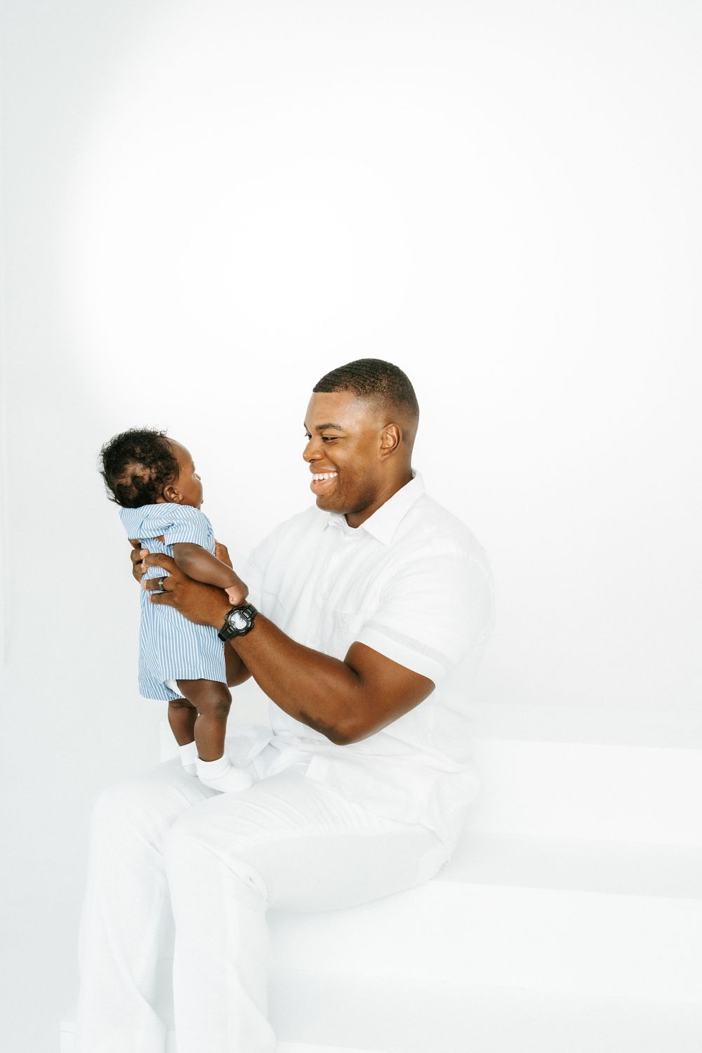 Smiling Black father holding his baby in a blue outfit against a white background.