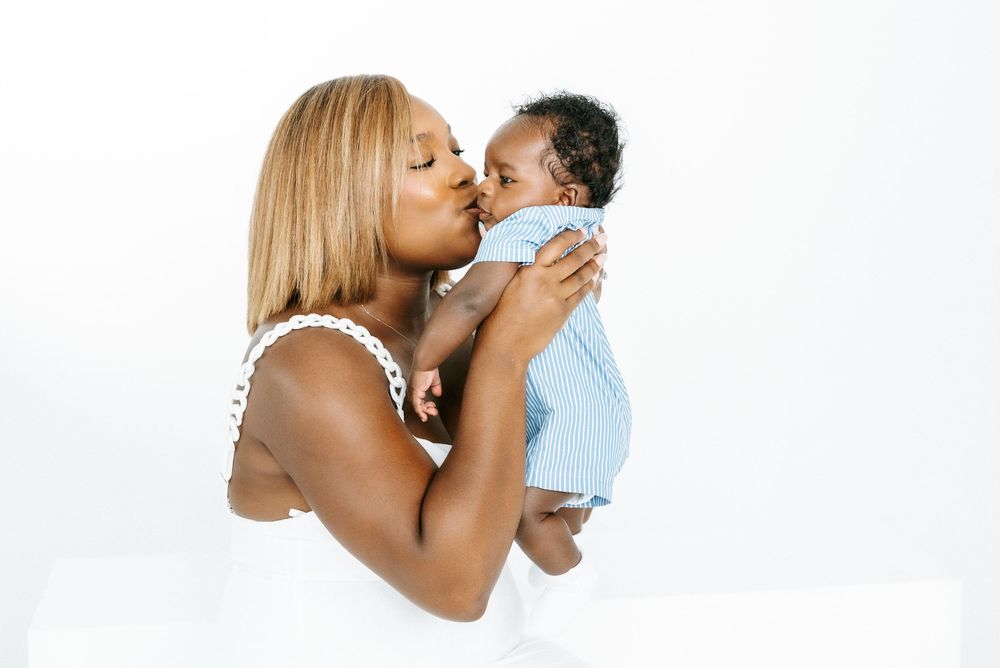 A loving Black mother kisses her newborn baby dressed in blue against a white background.
