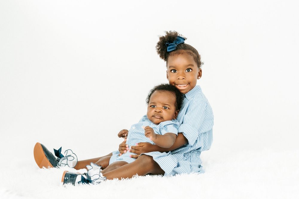 Two young siblings pose together on white background, older girl in blue dress hugging baby brother in white onesie.