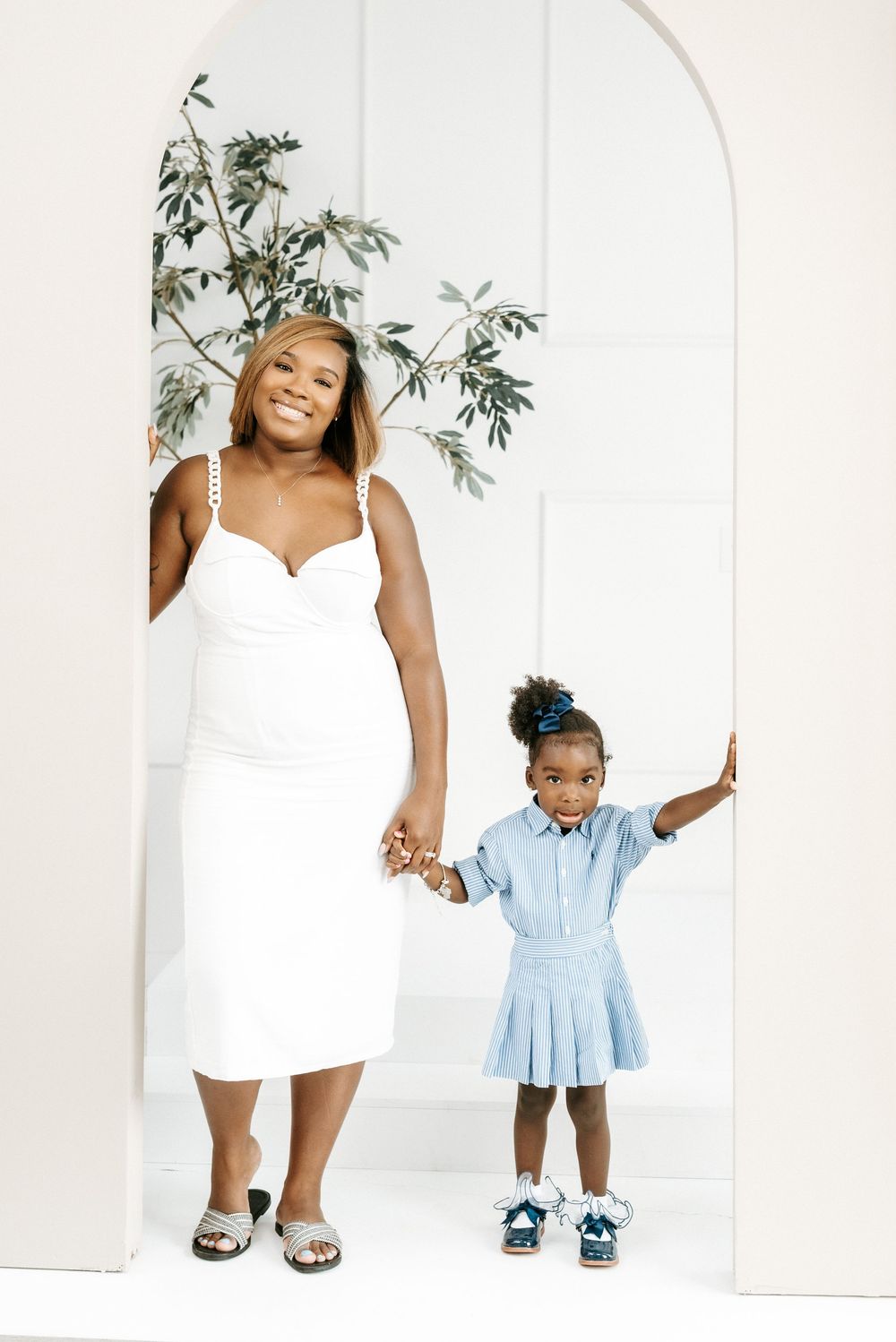 A pregnant Black woman in a white dress holds hands with a young girl in a blue dress standing in a white arch doorway.