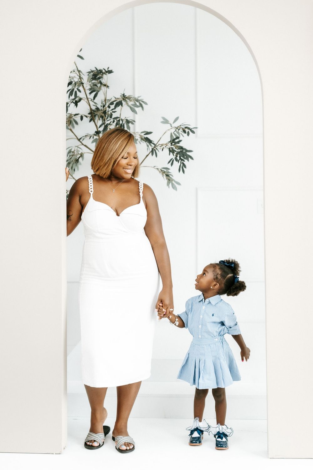 A woman in a white dress holds hands with a young girl in a blue dress standing in a white arched doorway.