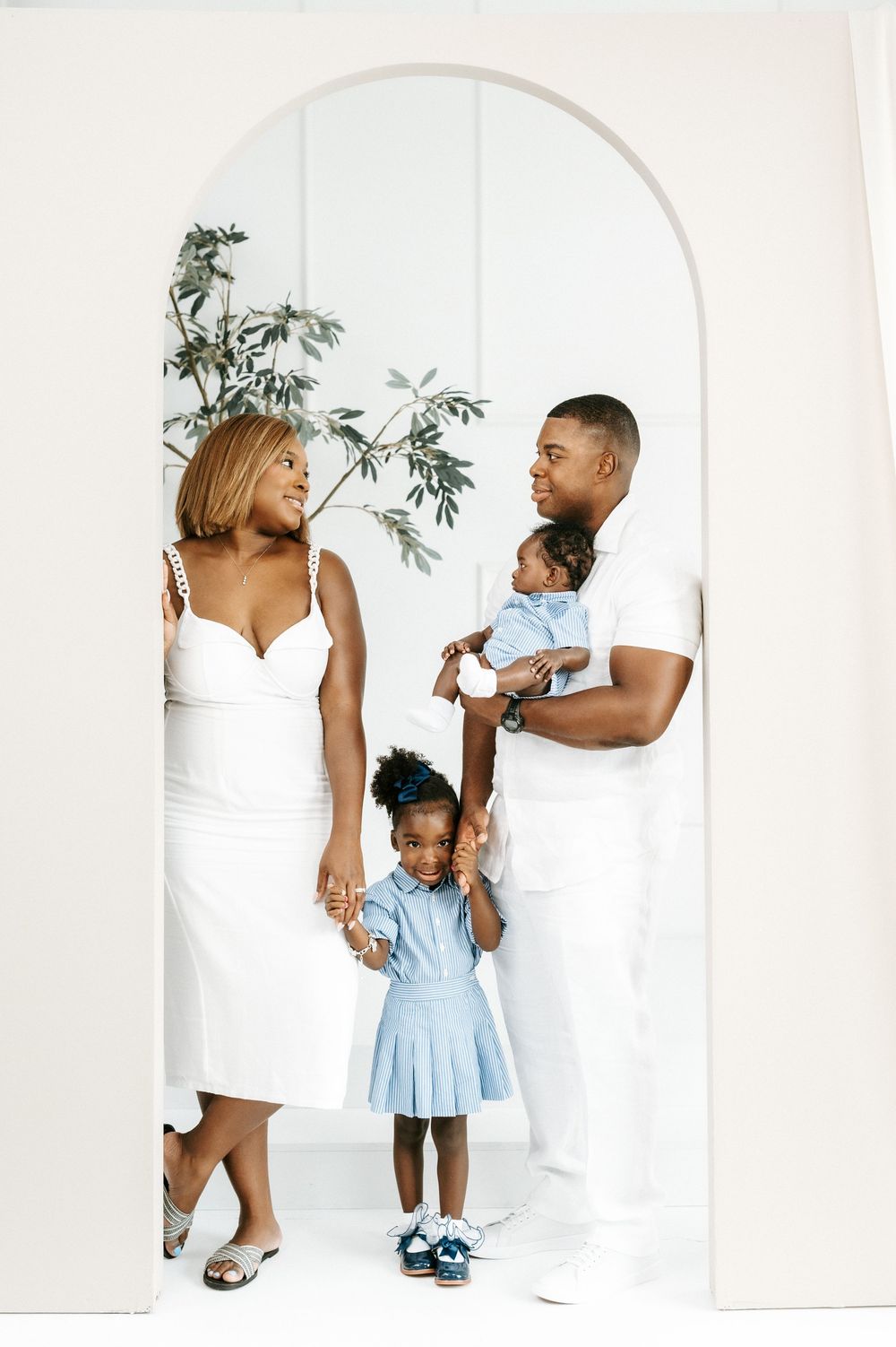 A family of four in white and blue poses in an arched doorway with greenery in a bright white studio.