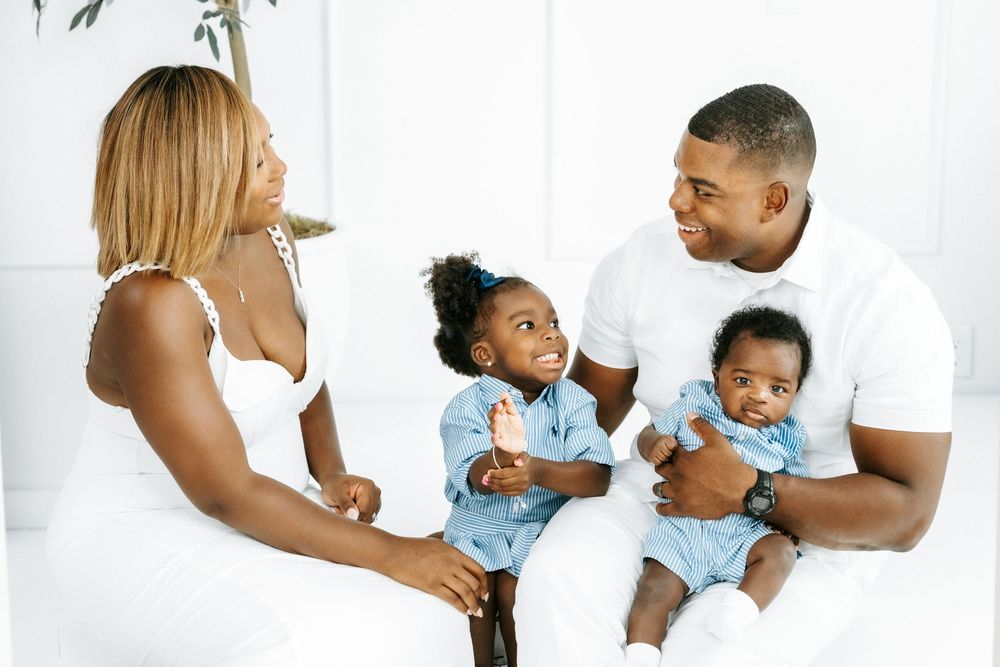 Happy Black family with mother, father, toddler girl, and baby boy dressed in white and blue outfits.