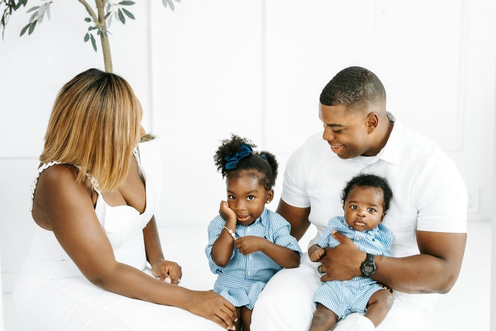 A happy Black family with two young children sitting together in a bright white room, dressed in matching outfits.