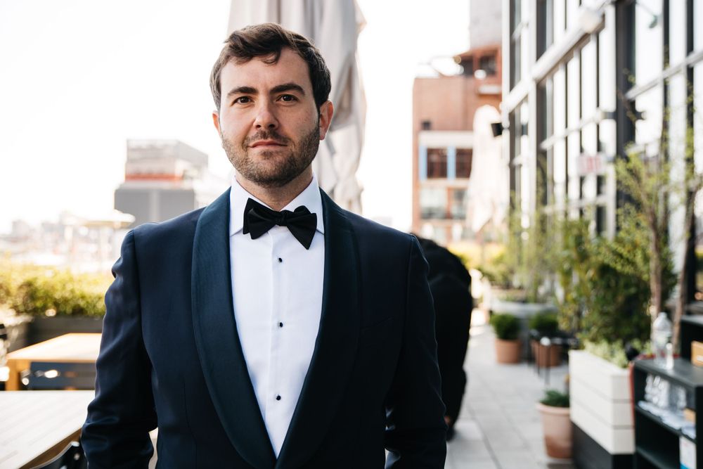 Dapper groom in navy tuxedo and bow tie standing on city street.