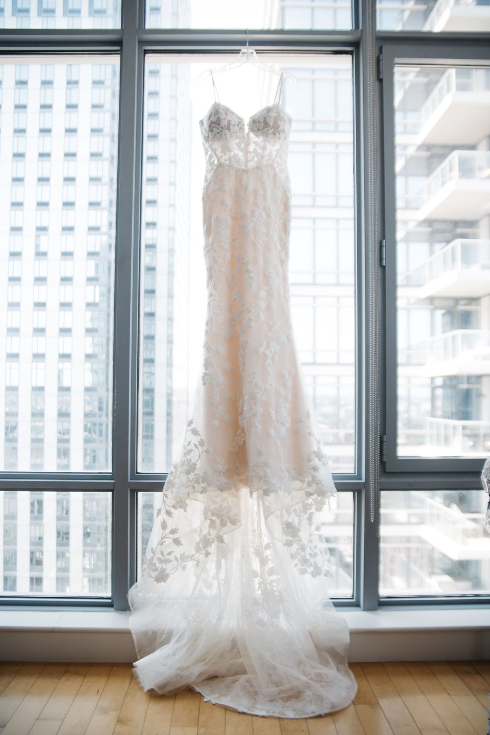 Delicate lace wedding gown hanging in front of floor-to-ceiling windows with city view.