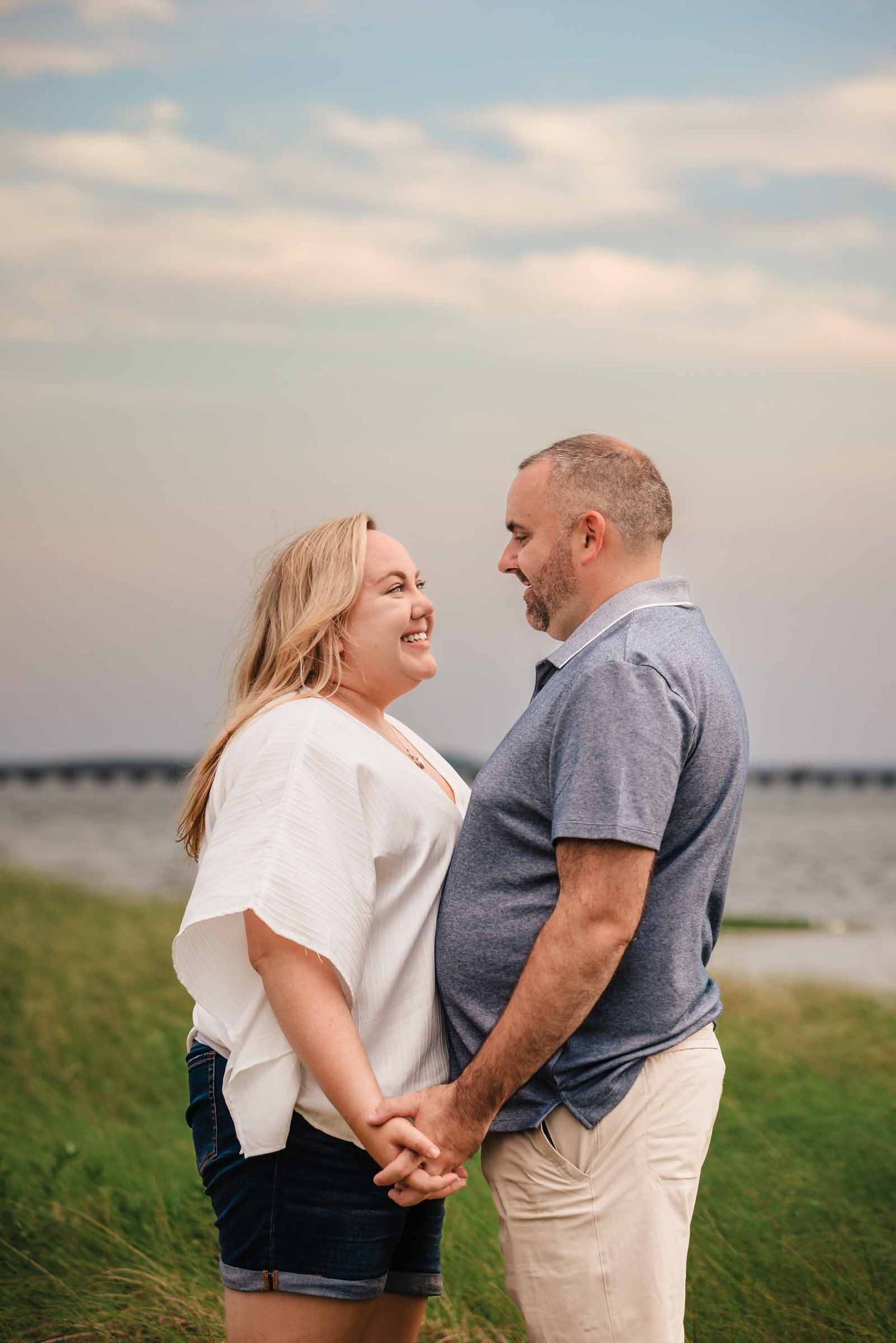 Couple holding hands and smiling at each other on a grassy waterfront at sunset.