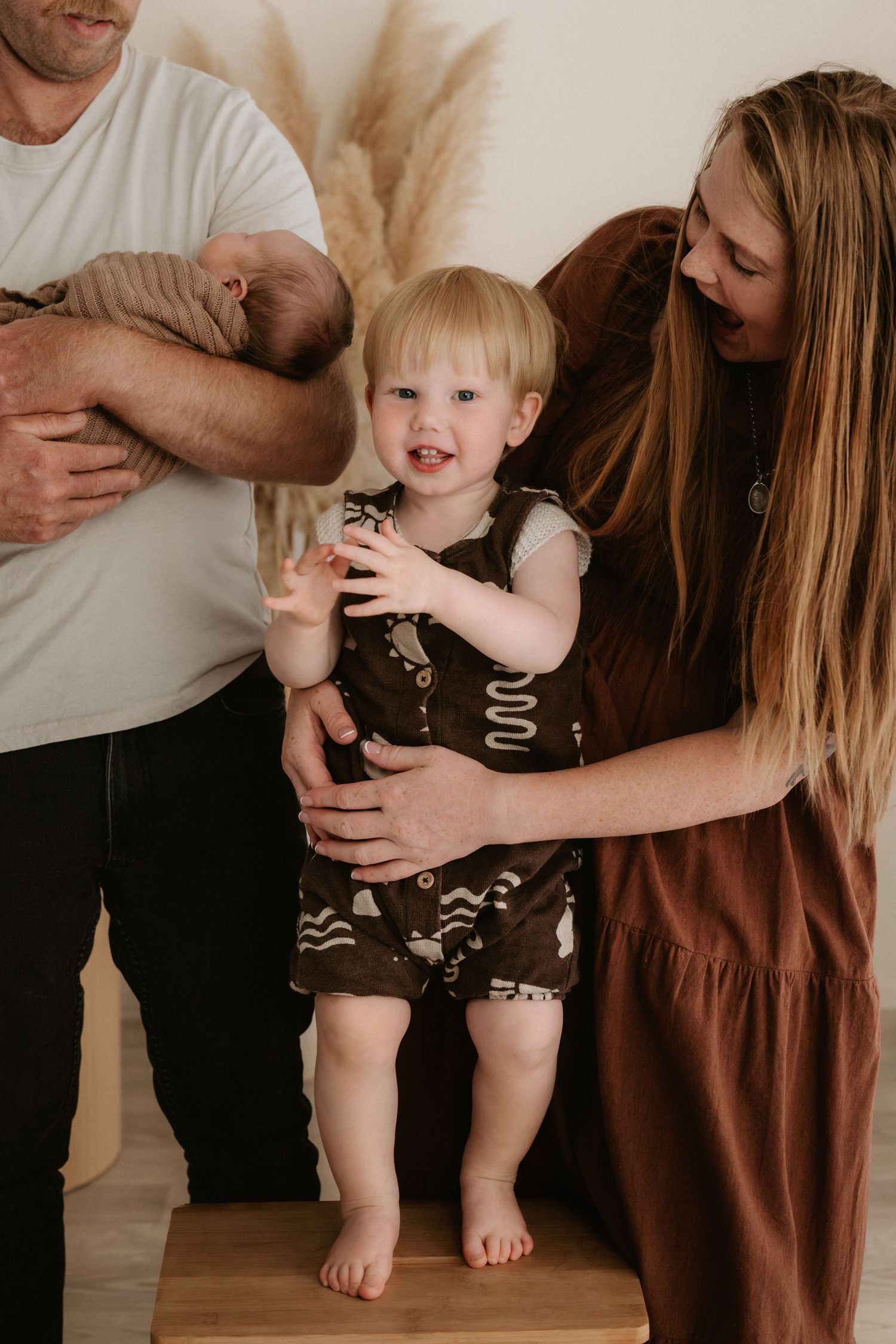 A toddler in a dark romper stands on a wooden block, supported by parents, one holding a newborn, in a bright studio.