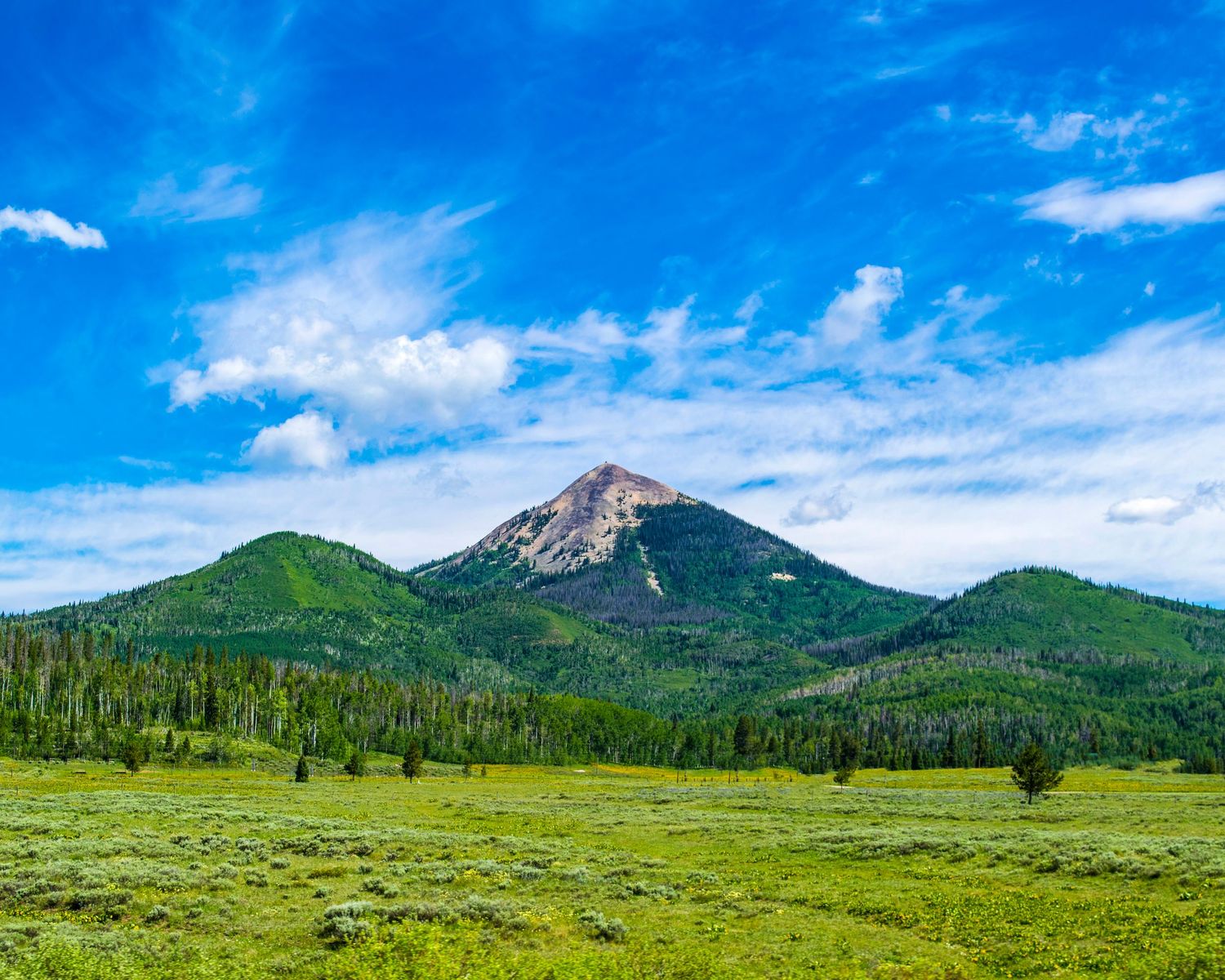 A scenic mountain peak rises above lush green meadows and forested slopes under a bright blue sky with white clouds.