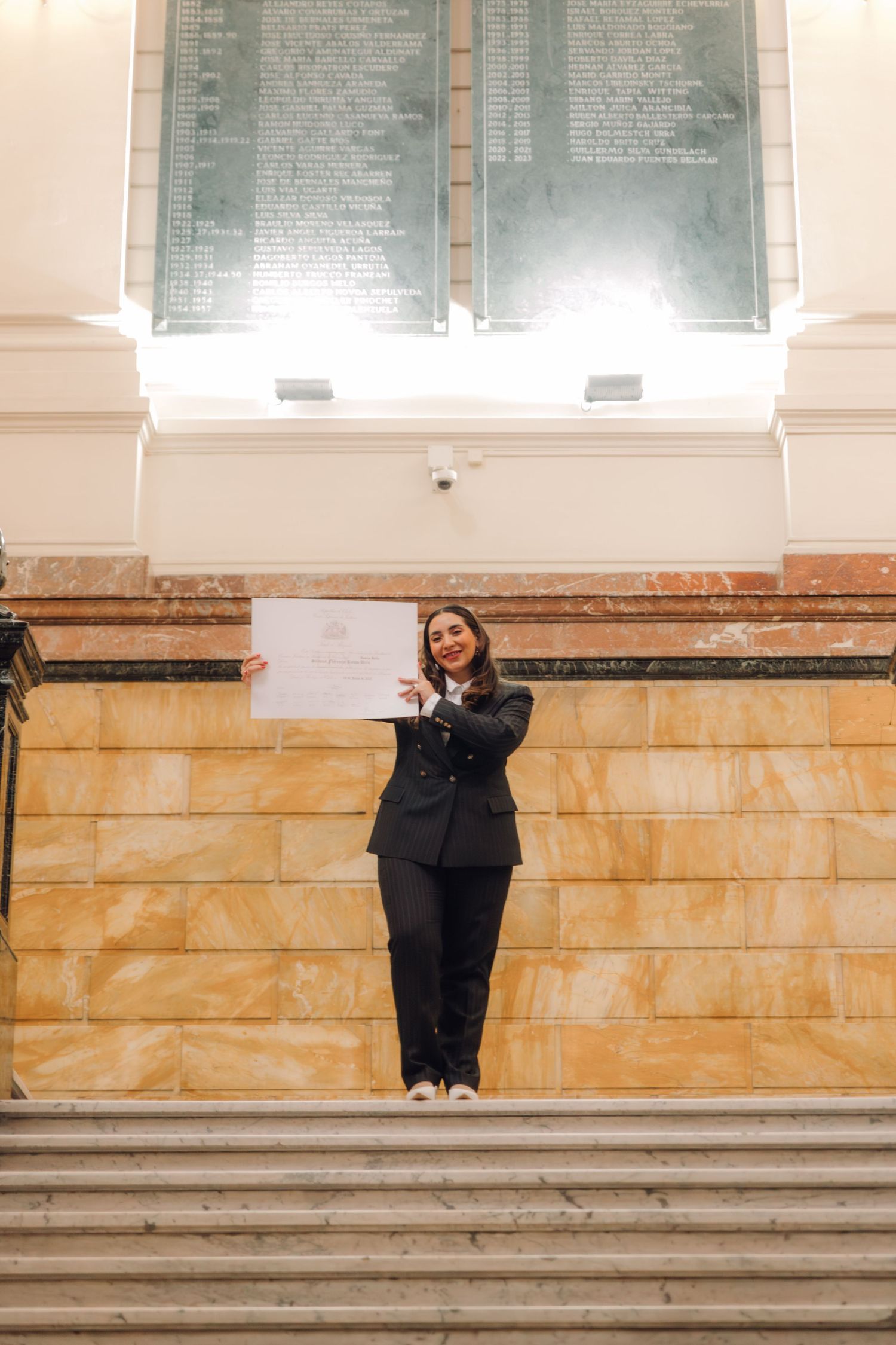 Smiling woman in black blazer holds diploma on marble staircase in classical building.