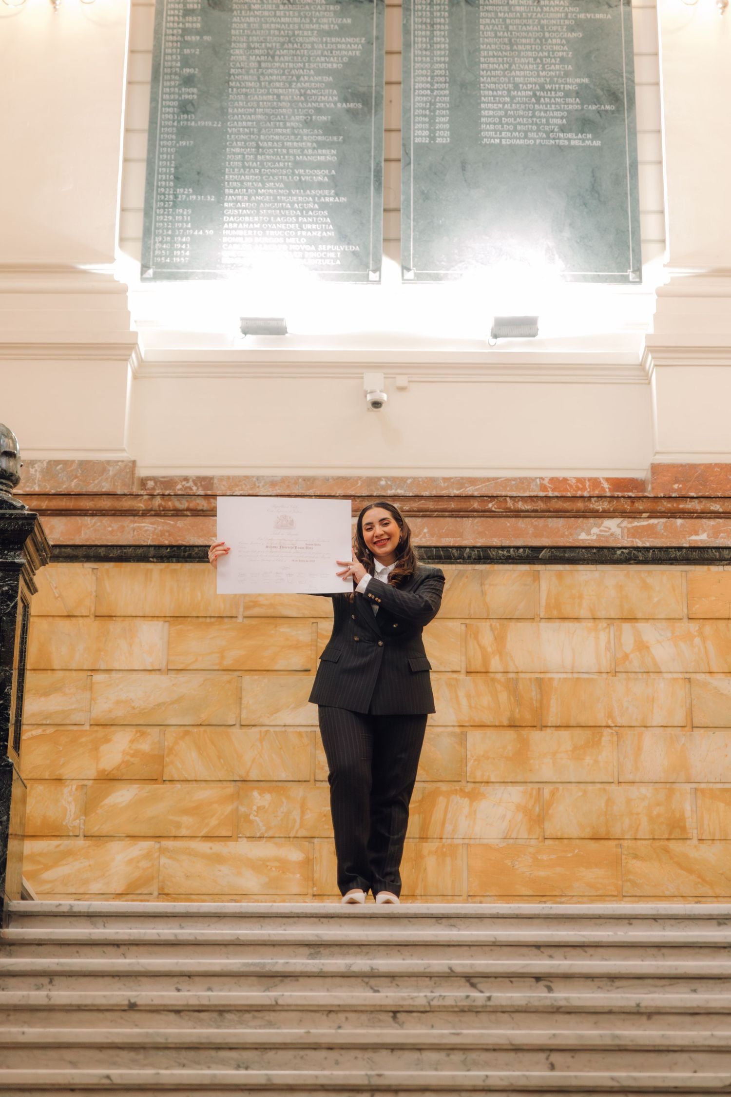 Woman in black suit displays certificate while standing on grand staircase in ornate building.