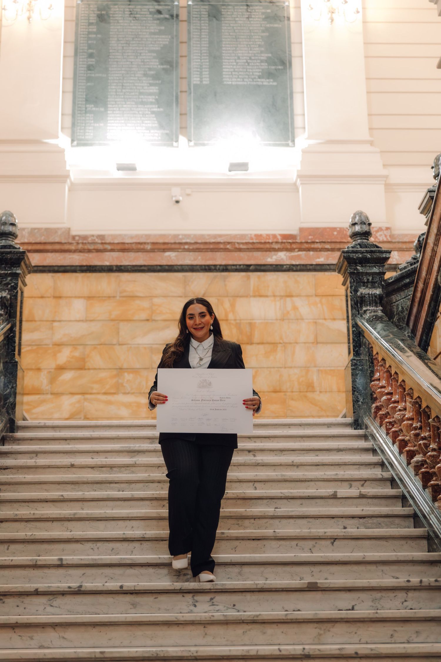 Graduate in black suit holds official certificate on stone staircase in historic building.