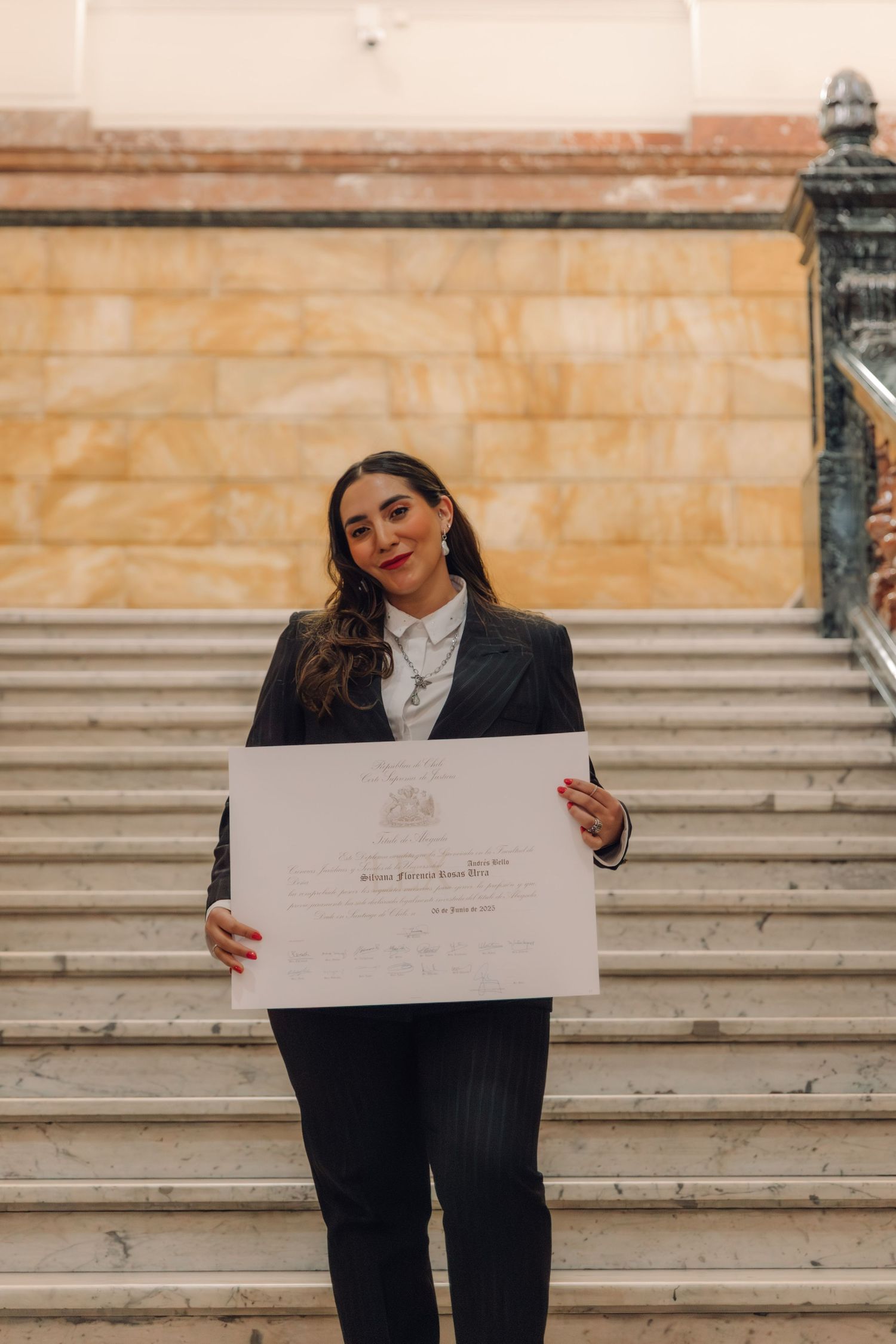 Woman in pinstripe suit proudly displays graduation certificate on outdoor stone staircase.