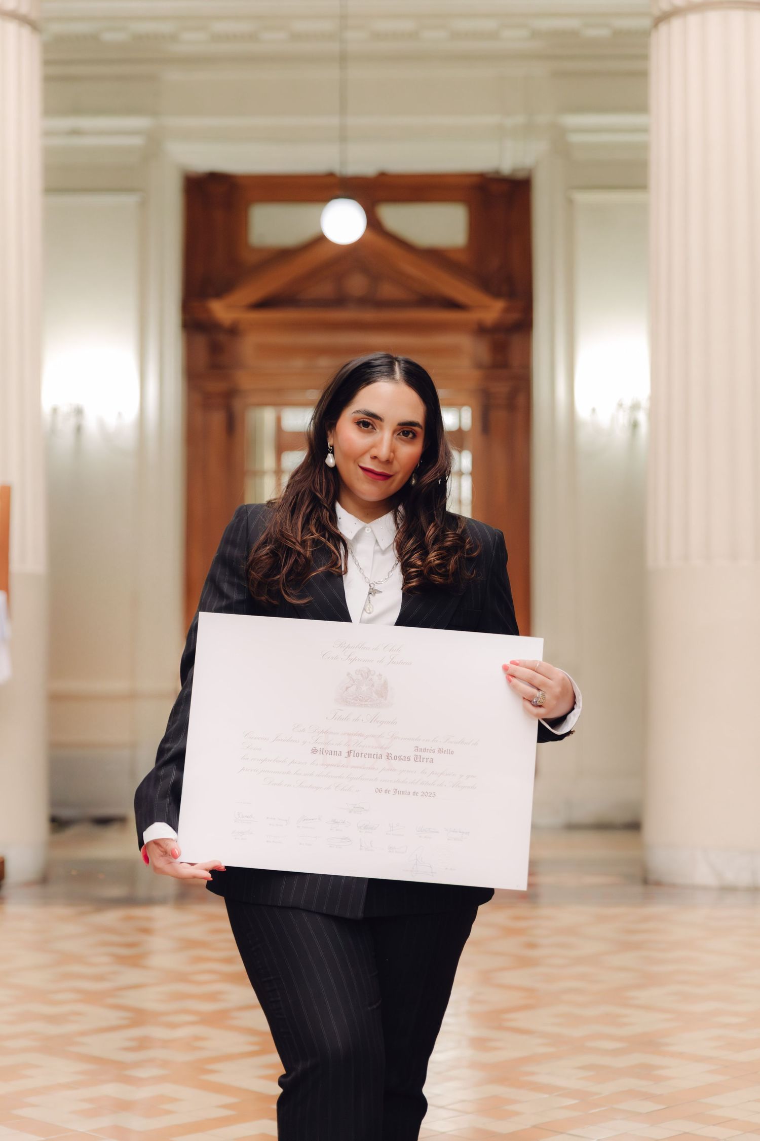 Woman in black pinstripe suit holds diploma certificate in elegant columned hallway.