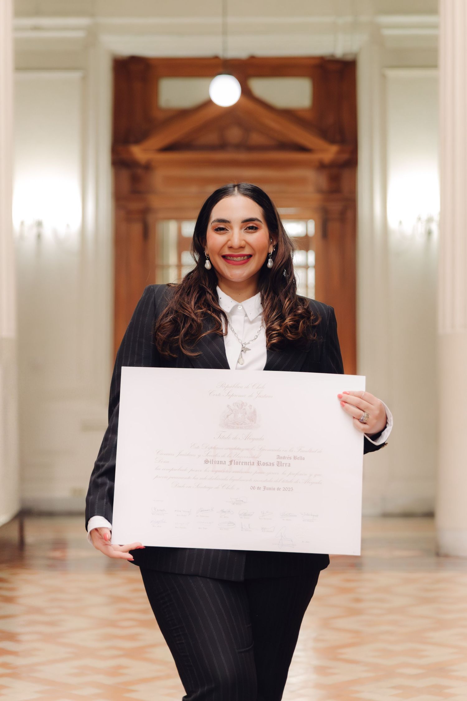 Smiling woman in pinstripe suit proudly holds large diploma in grand ceremonial hallway.