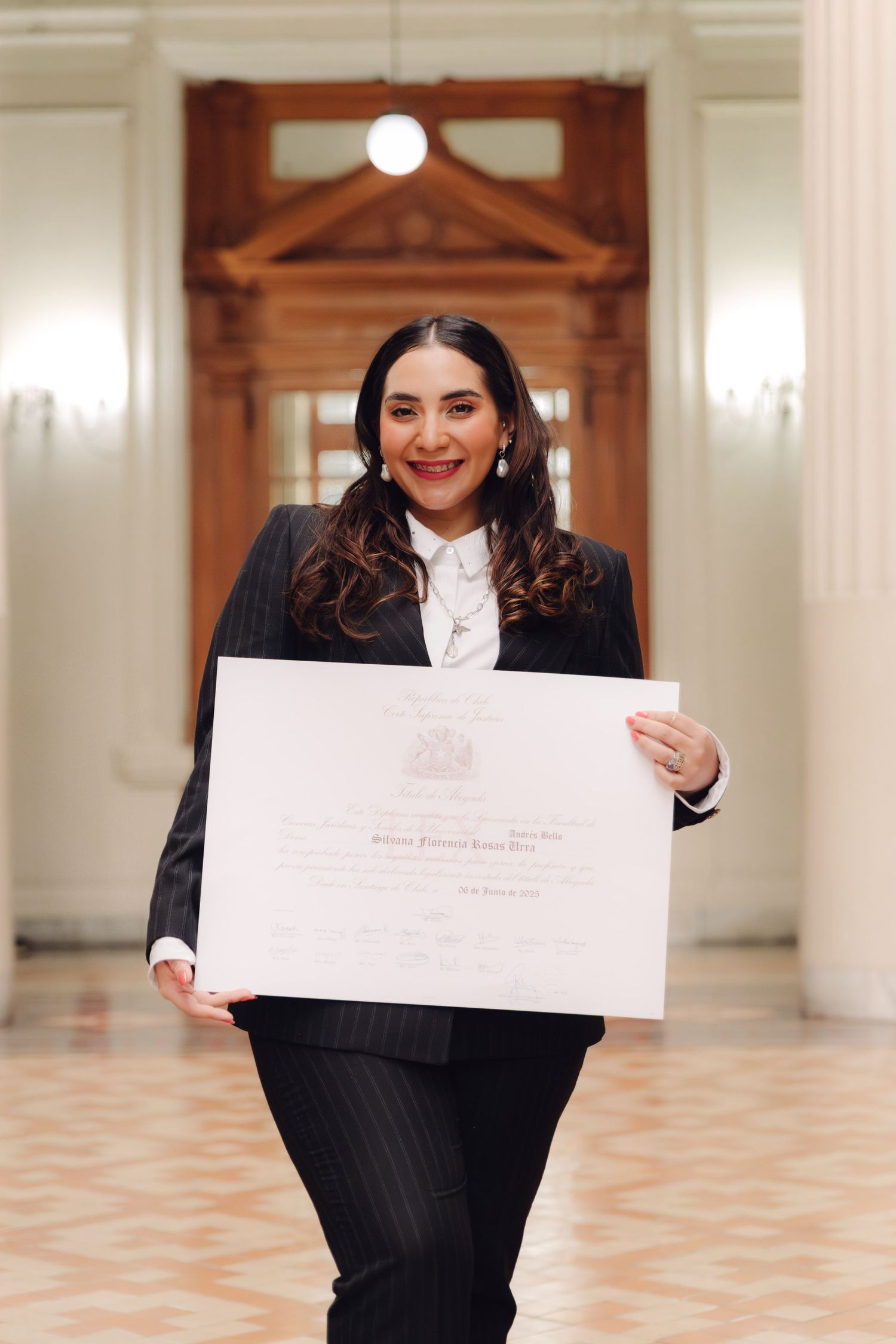 Woman in black pinstripe suit holds graduation certificate in elegant classical corridor.