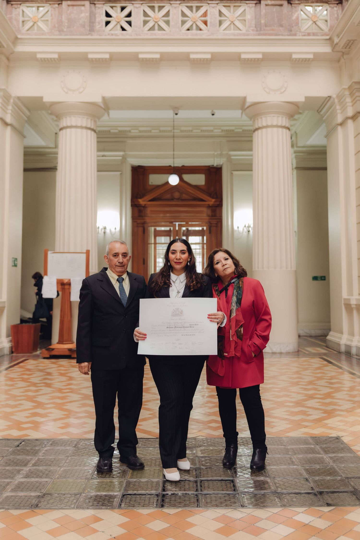 Graduate poses with parents holding diploma certificate in grand columned building foyer.
