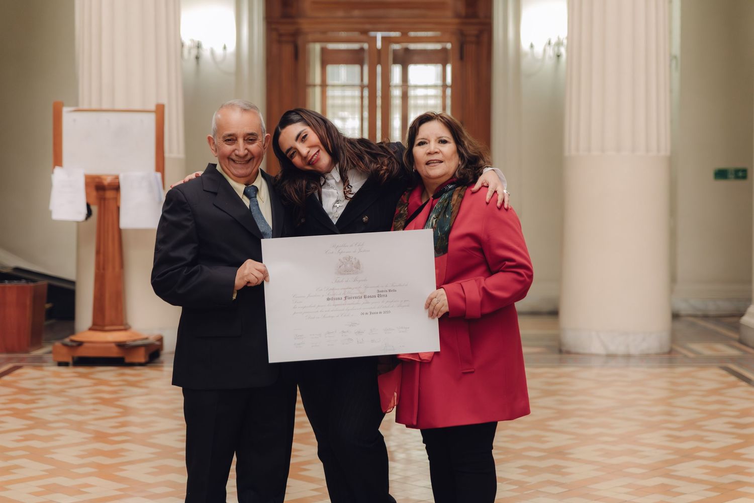 Three people proudly pose with a framed diploma certificate in an elegant hall.