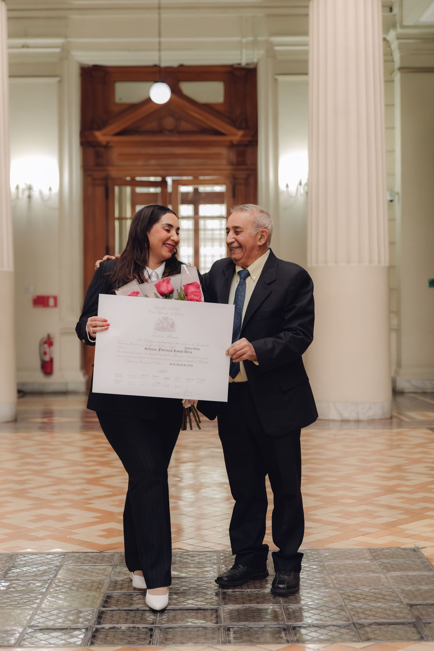 Graduate in black suit holds diploma and roses with older man in formal hall.