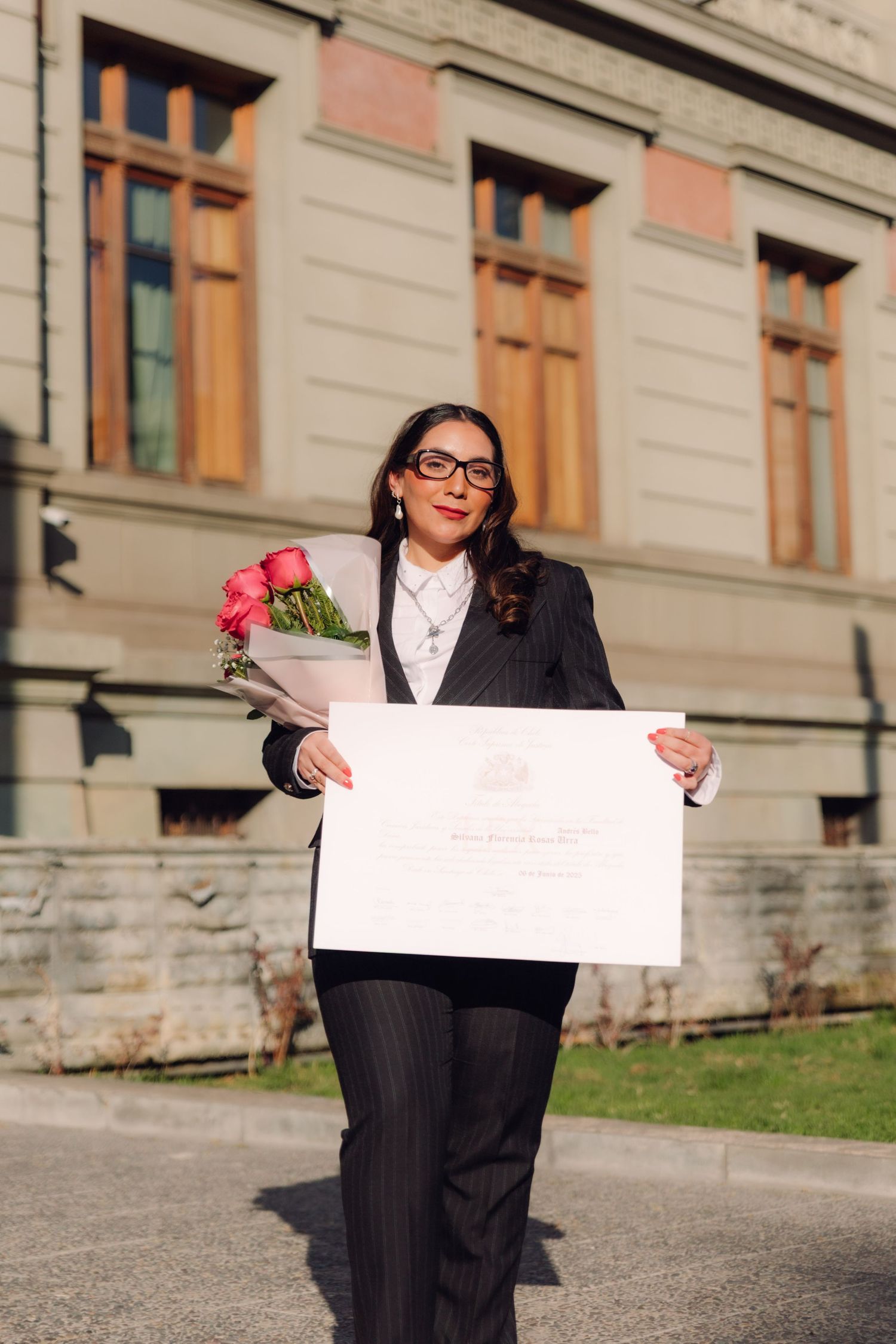 Graduate in black suit holds diploma and roses outside historic stone building.