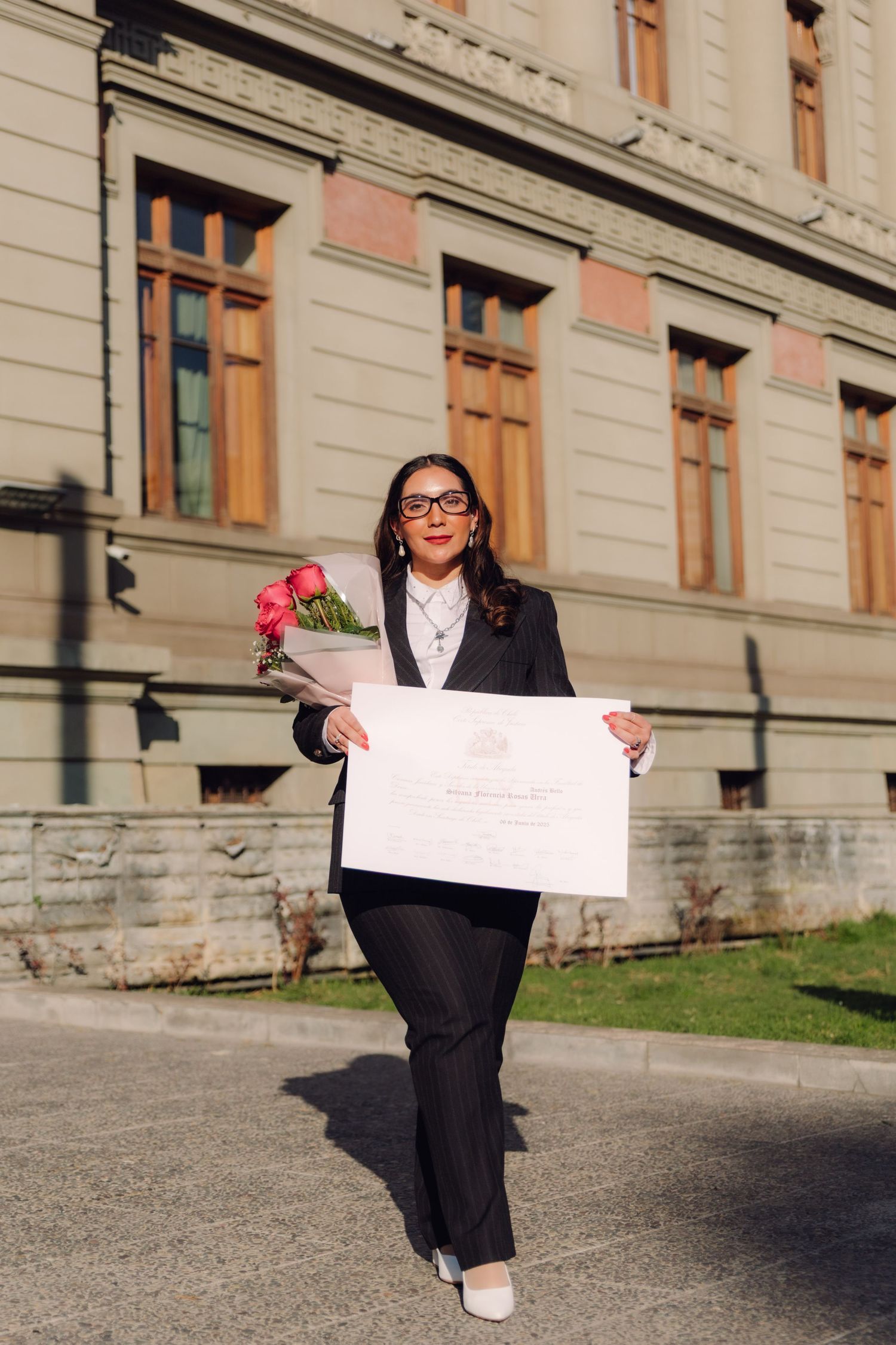 Woman in black suit holding diploma and pink roses outside historic building.