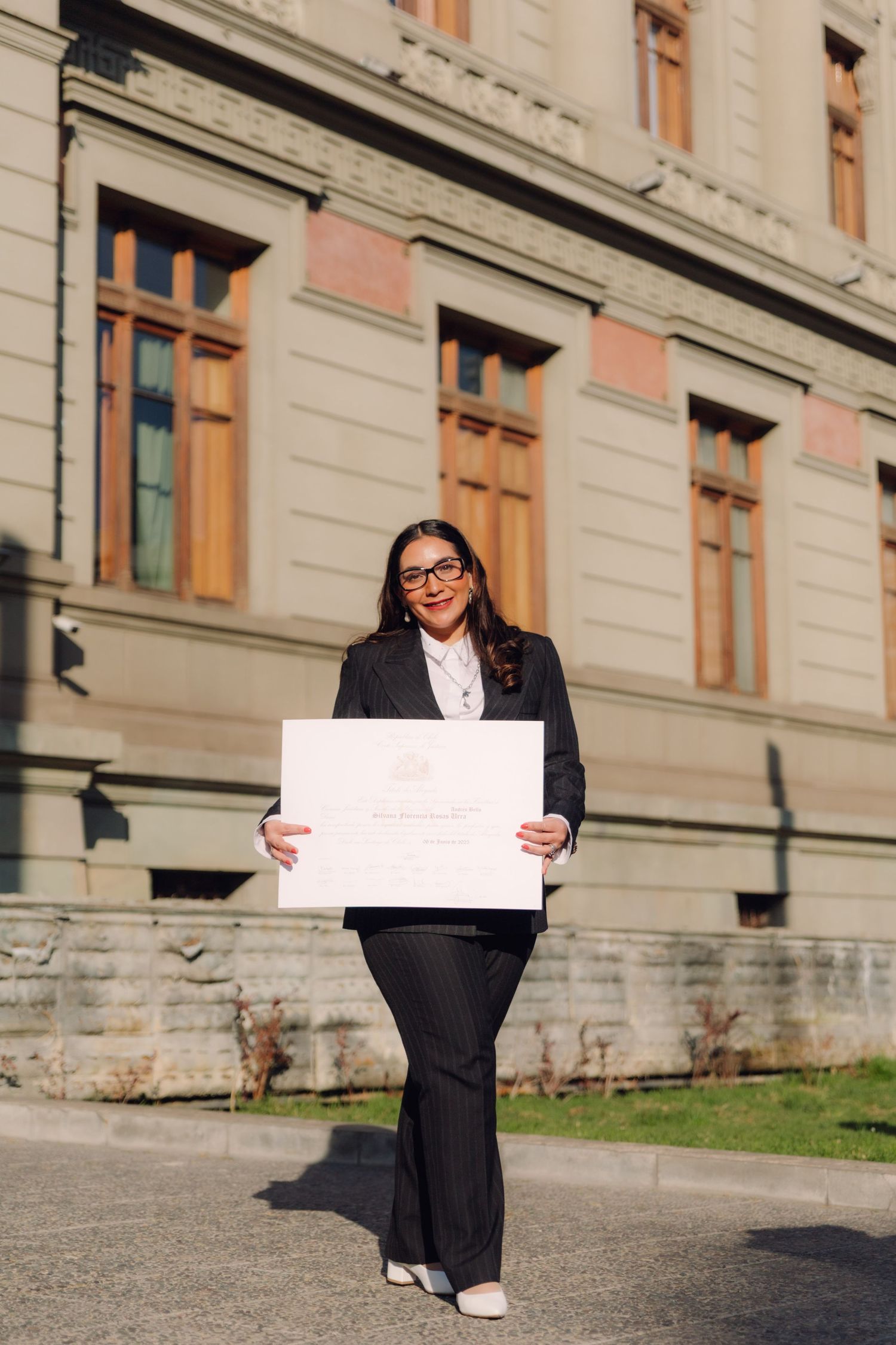 Smiling woman in black suit proudly displaying large diploma outside university building.