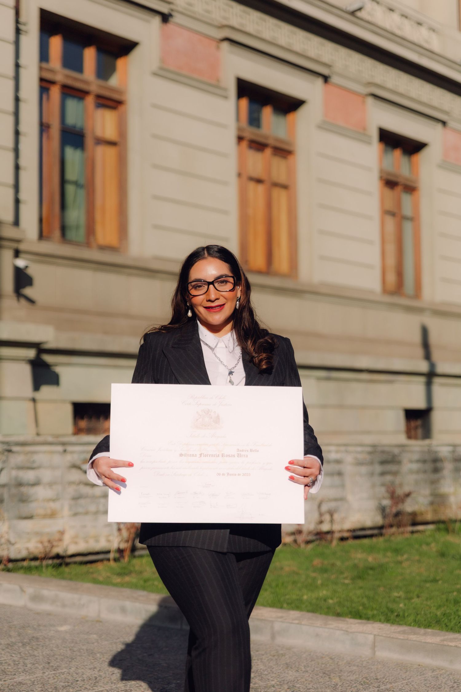 Woman in pinstripe suit holding official diploma certificate outside academic building.