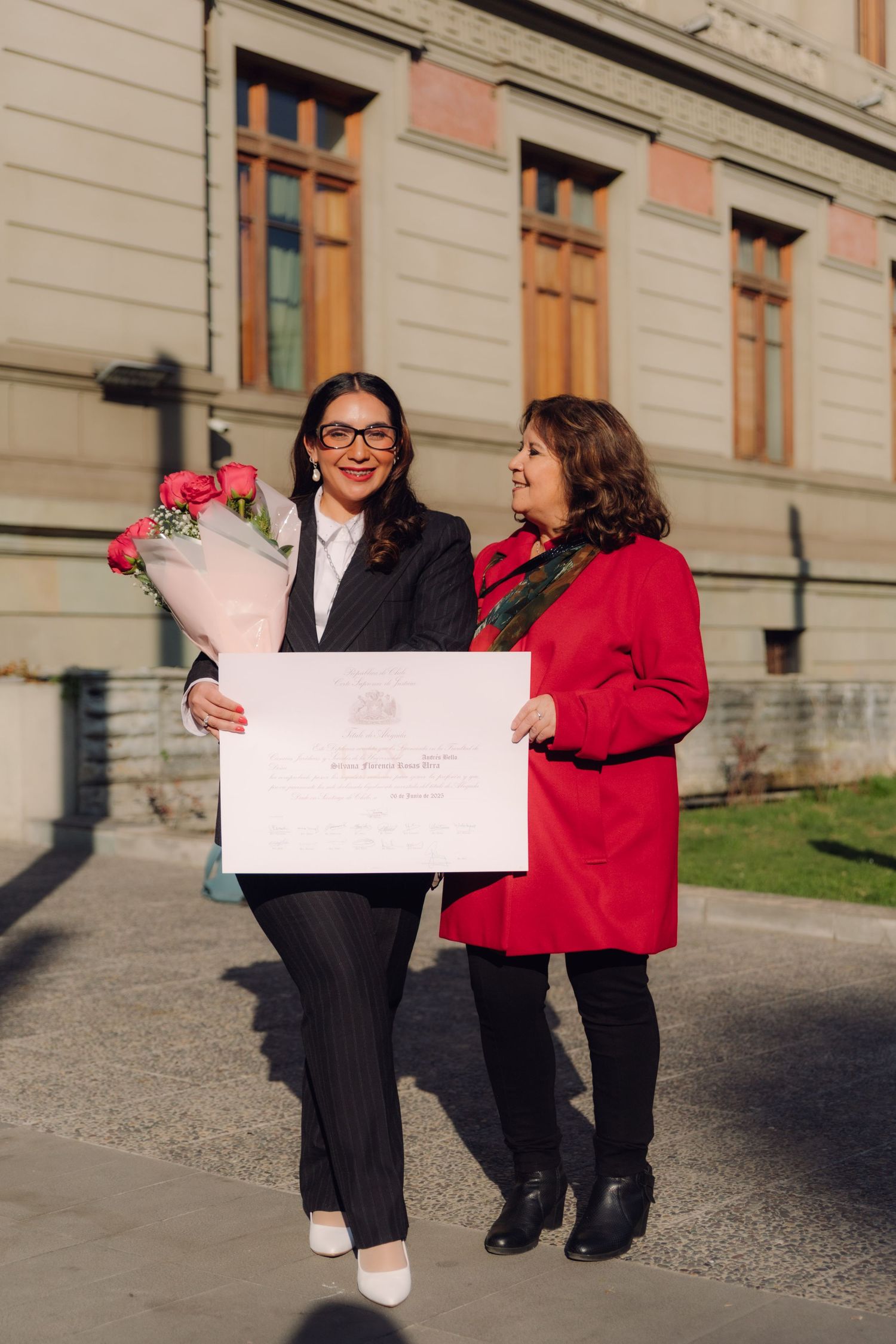 Two women sharing diploma and roses celebration outside historic building.