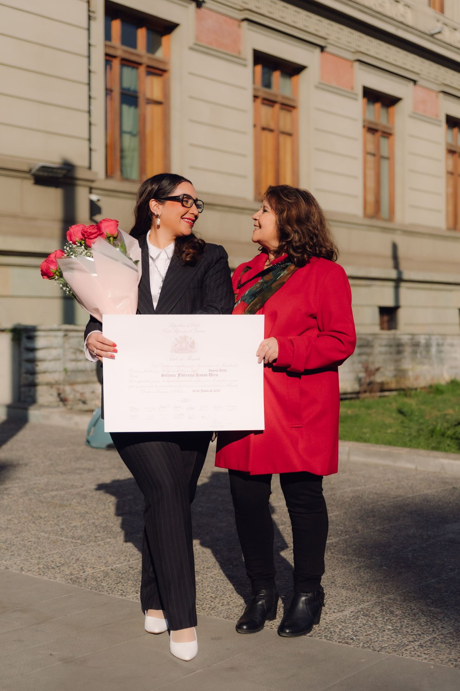 Two smiling women holding diploma and pink roses outside university building.